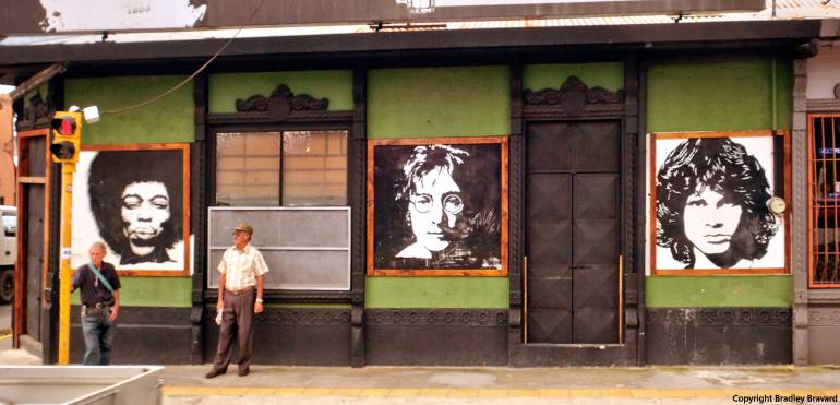 Two men standing on sidewalk and storefront with black and white portraits of Jimi Hendrix, John Lennon, and Jim Morrison